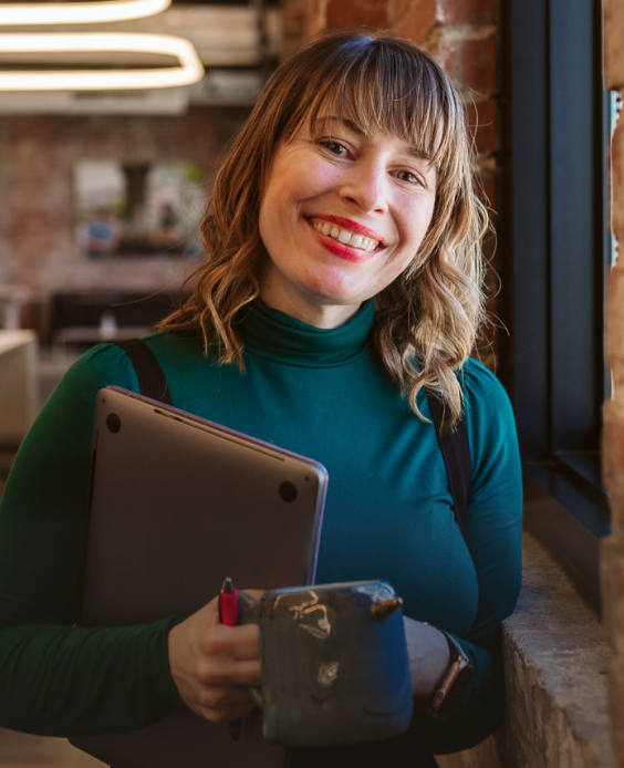 Woman standing next to a window smiling and holding a closed laptop and coffee mug