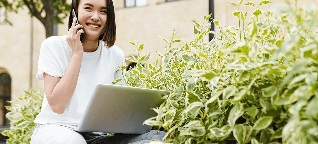 a woman works outside next to a bunch of greenery. she talks on the phone while holding a laptop