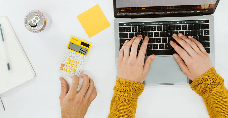 overhead shot of arms wearing yellow sleeves are poised above a laptop keyboard, ready to type. to the left of the computer, a calculator, post it notes, a notebook, and another person's hand are visible