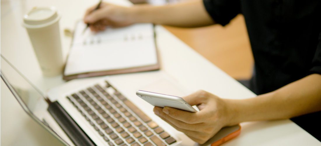 Photo of a person working on laptop and phone while writing in notebook