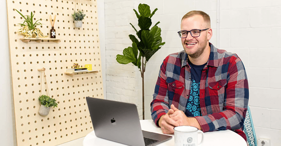 a man in a red and blue flannel shirt sits in front of his laptop at a table in a white office space