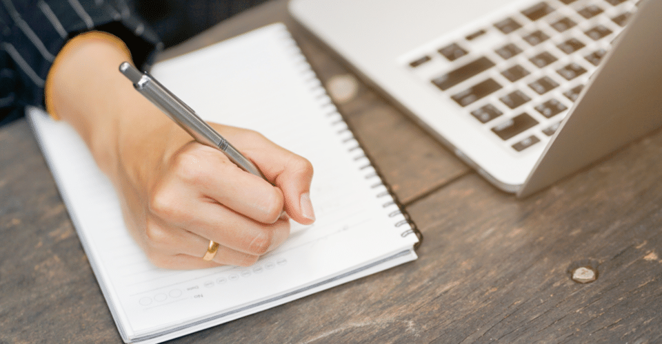 person writing in notebook next to macbook